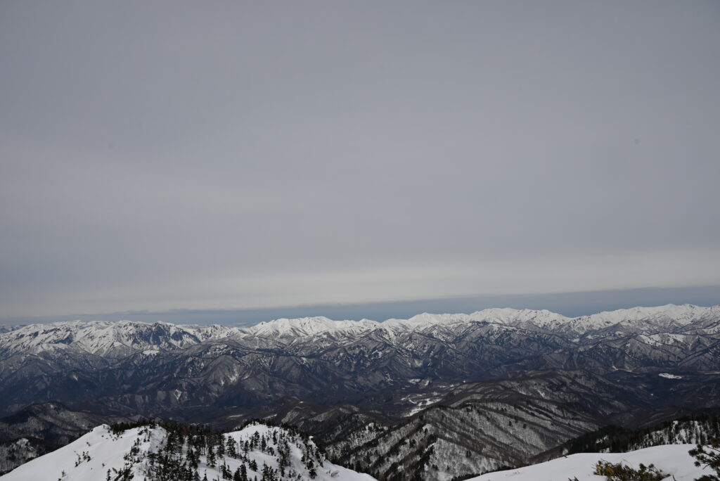 山頂から西側の山々を望む　左の黒っぽい山肌（一ノ倉沢）を持つのは谷川岳　その隣に、朝日岳、巻機山が連なる。